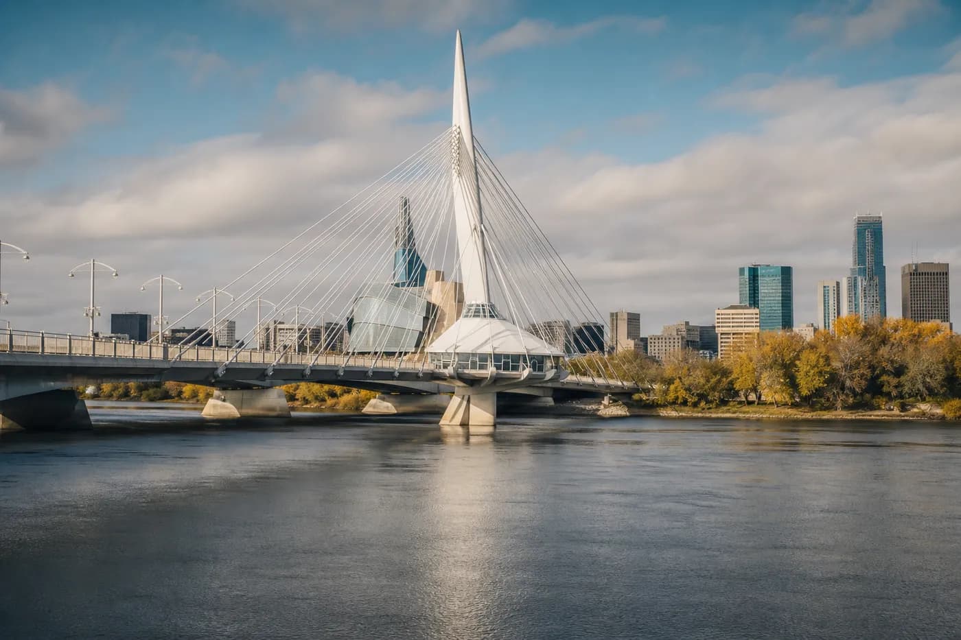 Cable-stayed pedestrian bridge over the Red River with the Winnipeg skyline and autumn trees behind it, Manitoba