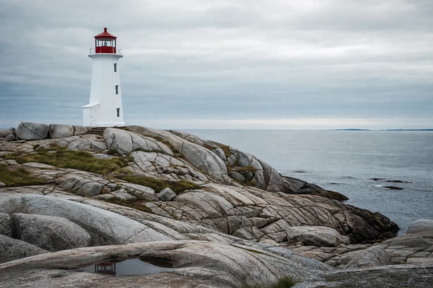White and red Atlantic lighthouse standing on wave-worn granite boulders above a calm grey-blue sea, Nova Scotia